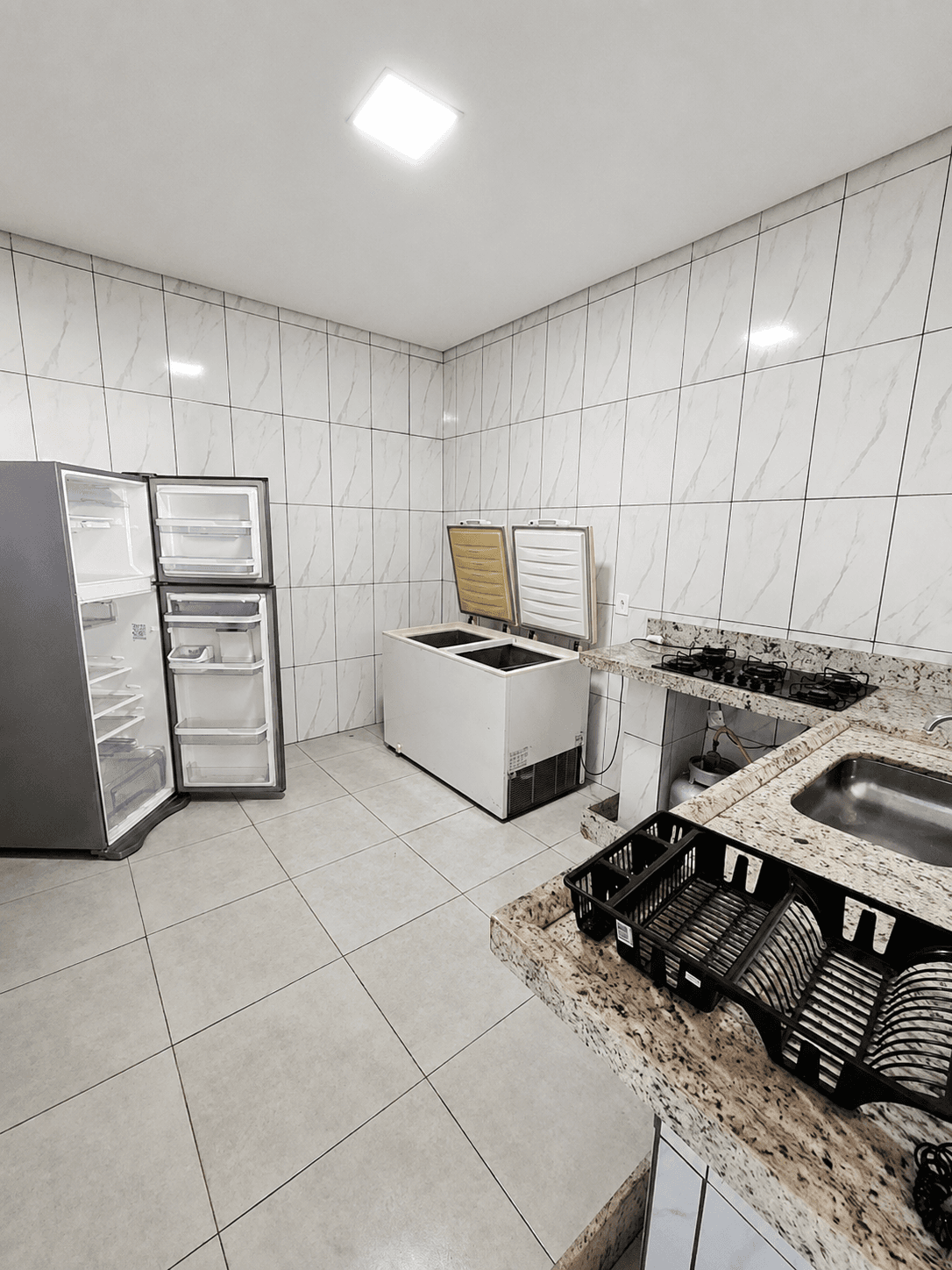 Cozinha de apoio da área gourmet, com geladeira aberta, dois freezers e cooktop com pia ao fundo.